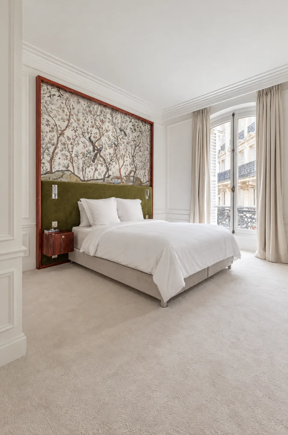 Chambre lumineuse avec tête de lit végétale et fenêtre sur une façade haussmannienne